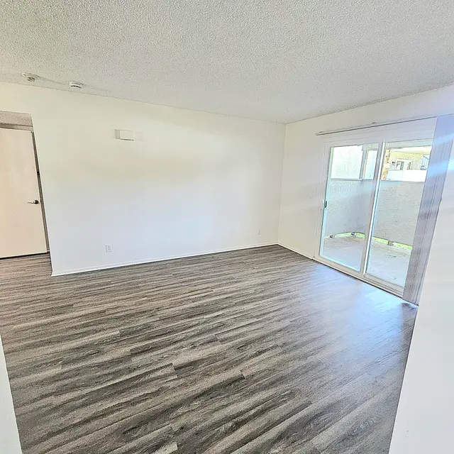 A spacious, empty room with light-colored walls and dark wood-like flooring. A sliding glass door leads to a small outdoor area. A door is visible on the left side.