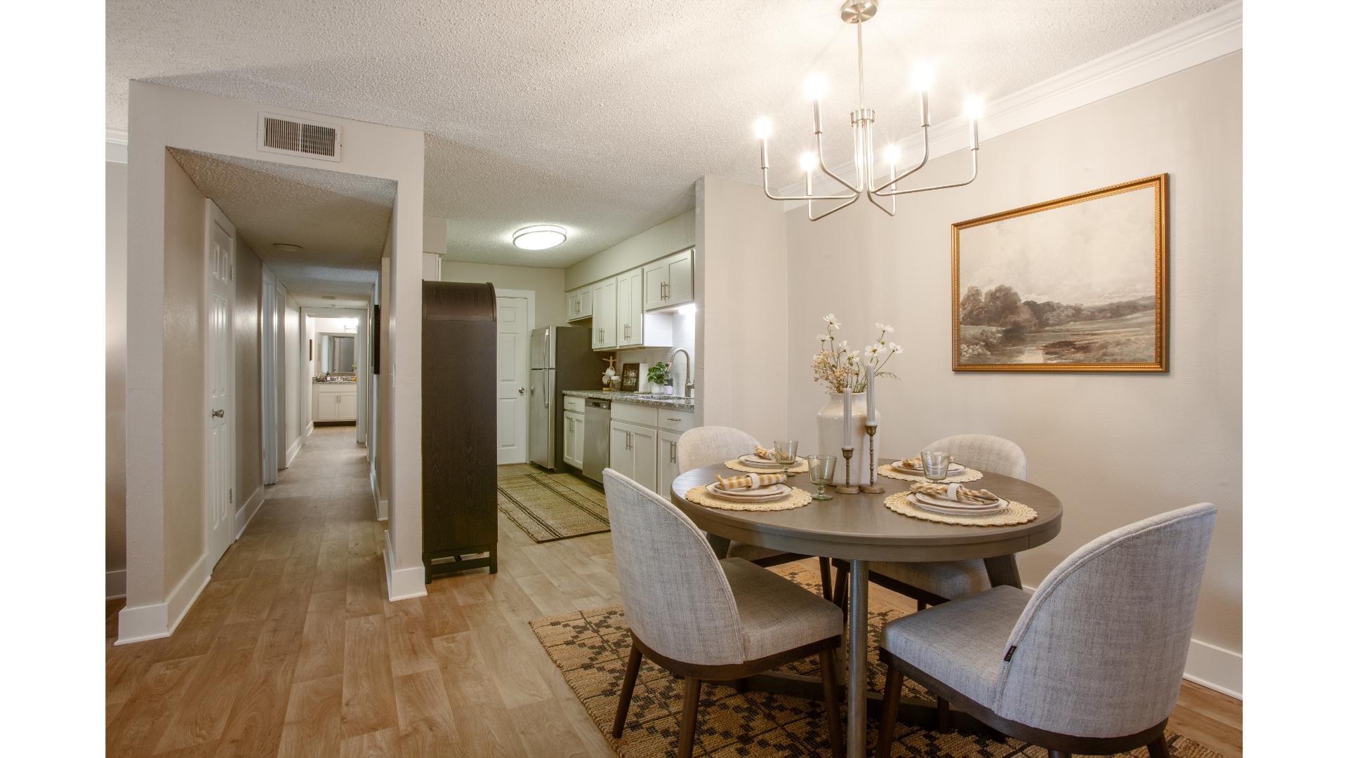 Stylish Dining Area and Kitchen A dining area featuring a round table set for four with gray upholstered chairs, a chandelier above, and a painting on the wall. In the background, a kitchen is visible, and a hallway leads out of the frame.