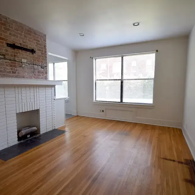 A vacant living room featuring a large window, wooden floors, and a brick fireplace.