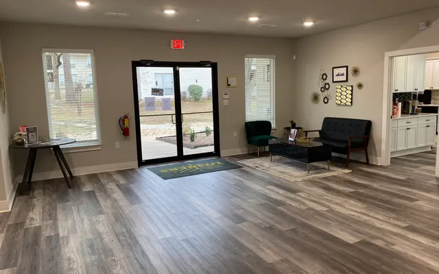 A spacious lobby area featuring a welcoming entrance with glass doors, wooden flooring, and stylish furniture including a dark couch and a wooden armchair. Natural light enters through two windows, enhancing the bright ambiance.