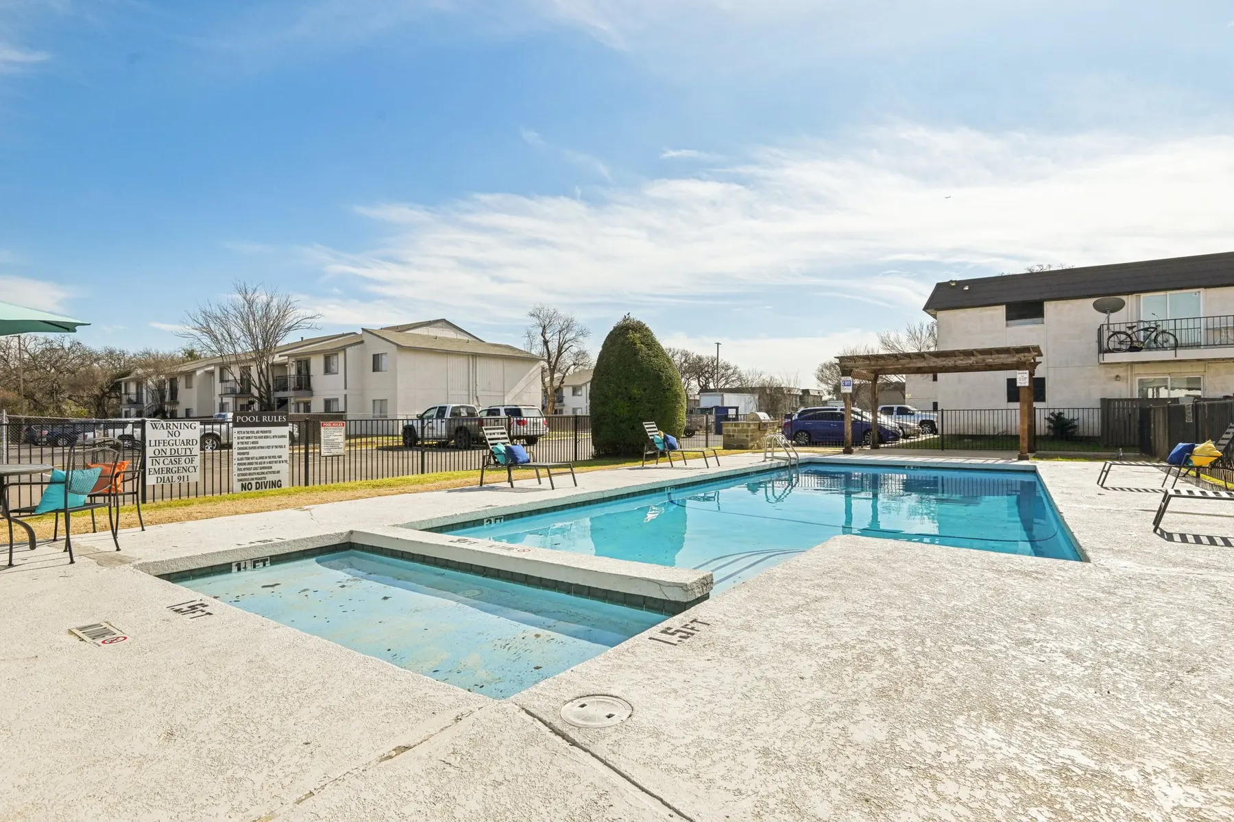 A swimming pool area with a main pool and a smaller spa, surrounded by lounge chairs and a fence, with residential buildings in the background and a clear blue sky overhead.