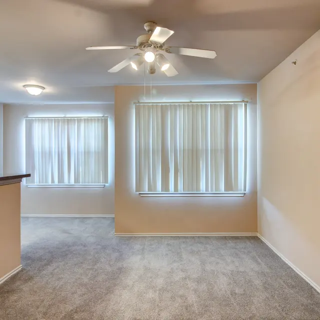 Spacious Living Room with Natural Light A bright and spacious living room featuring beige walls and light gray carpet. Two large windows with white curtains allow natural light to fill the room. A ceiling fan is visible, and a half wall separates the living area from another room.