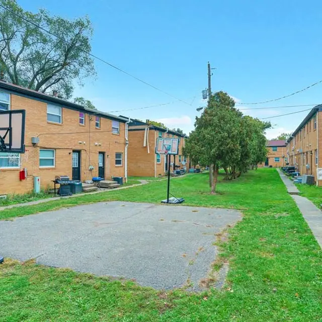 A view of an outdoor basketball court located between two rows of apartment buildings. The court is on an open grassy area with trees in the background, featuring basketball hoops but no players visible.