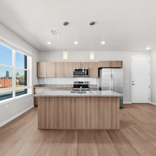 A spacious modern kitchen featuring wooden cabinets, stainless steel appliances, and a large central island with granite countertop, illuminated by pendant lights. Large windows allow natural light to flood the area, and the flooring is made of light-colored wood.