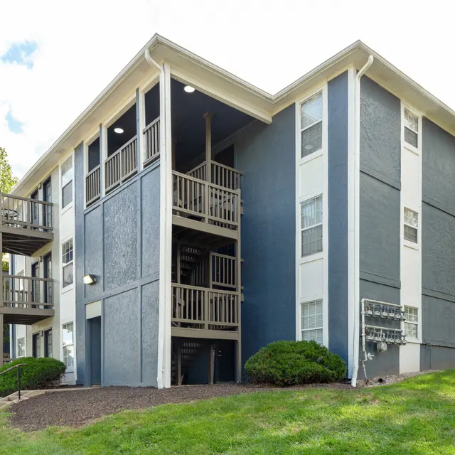 Exterior view of a modern apartment building with three levels and balconies, surrounded by greenery.