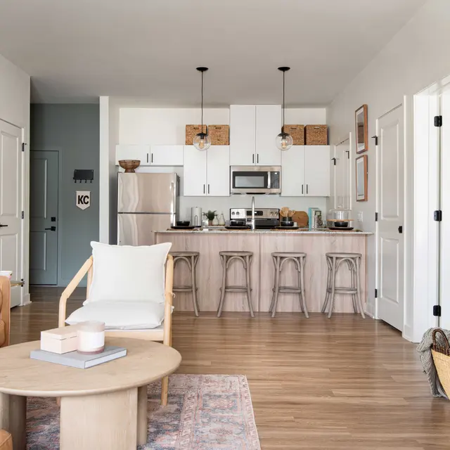 A spacious living room with a kitchen area featuring white cabinetry, a wooden dining table, and a cozy sofa. The decor is minimalistic with natural light illuminating the space.