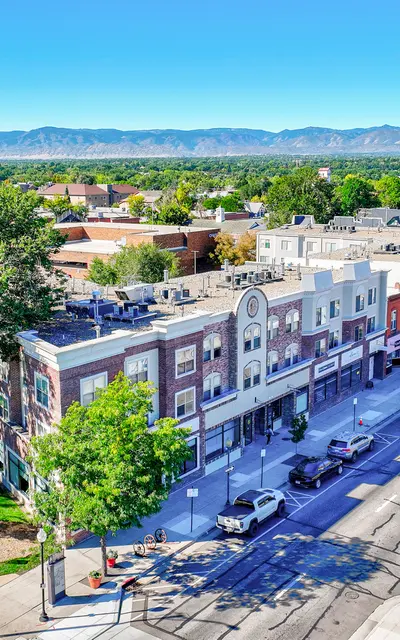 Aerial view of a downtown street lined with buildings and trees, showcasing shops and parked cars, with mountains in the background under a clear blue sky.