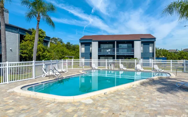 Apartment Complex Pool Area A clear, inviting swimming pool surrounded by a paved area, with a white fence and lounge chairs, under a bright blue sky with a few clouds.