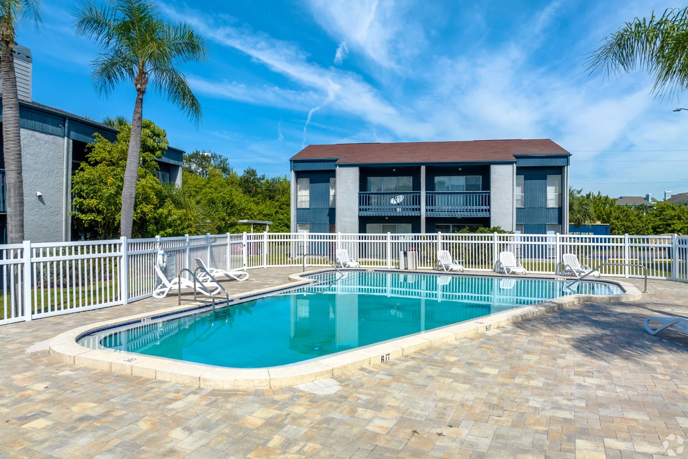 A clear, inviting swimming pool surrounded by a paved area, with a white fence and lounge chairs, under a bright blue sky with a few clouds.