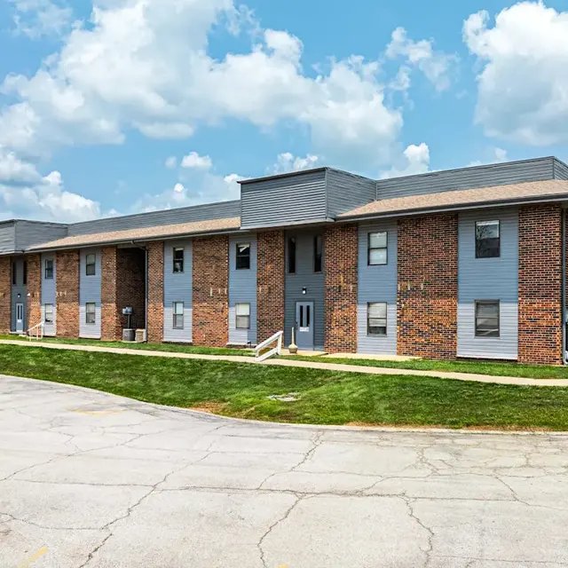 A modern apartment building featuring a mix of brick and gray siding, set against a bright blue sky with fluffy white clouds. The building has multiple entrance doors and large windows. Green grass surrounds the structure, and a curved driveway leads up to it.
