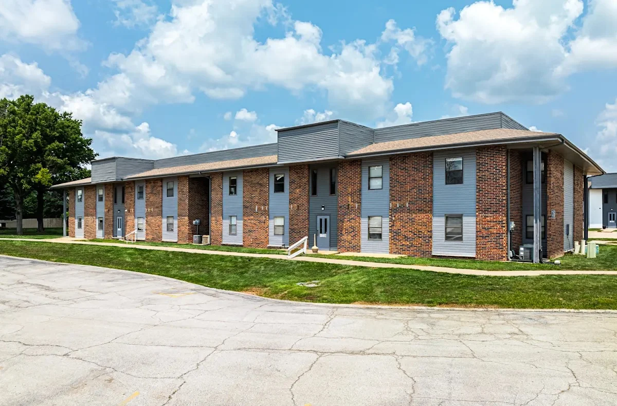 A modern apartment building featuring a mix of brick and gray siding, set against a bright blue sky with fluffy white clouds. The building has multiple entrance doors and large windows. Green grass surrounds the structure, and a curved driveway leads up to it.