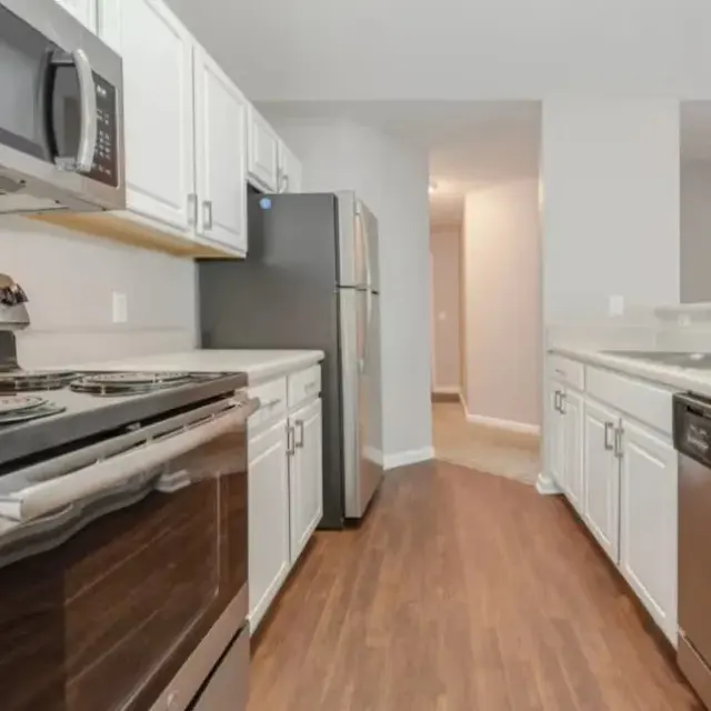 A modern kitchen with white cabinets, stainless steel appliances, and a wooden floor. The kitchen features a stove, microwave, refrigerator, and dishwasher, with a bright and spacious layout.