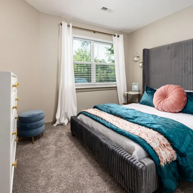 A cozy bedroom with a grey bed, teal accents, a large pink pillow, a white dresser, and natural light from a window.