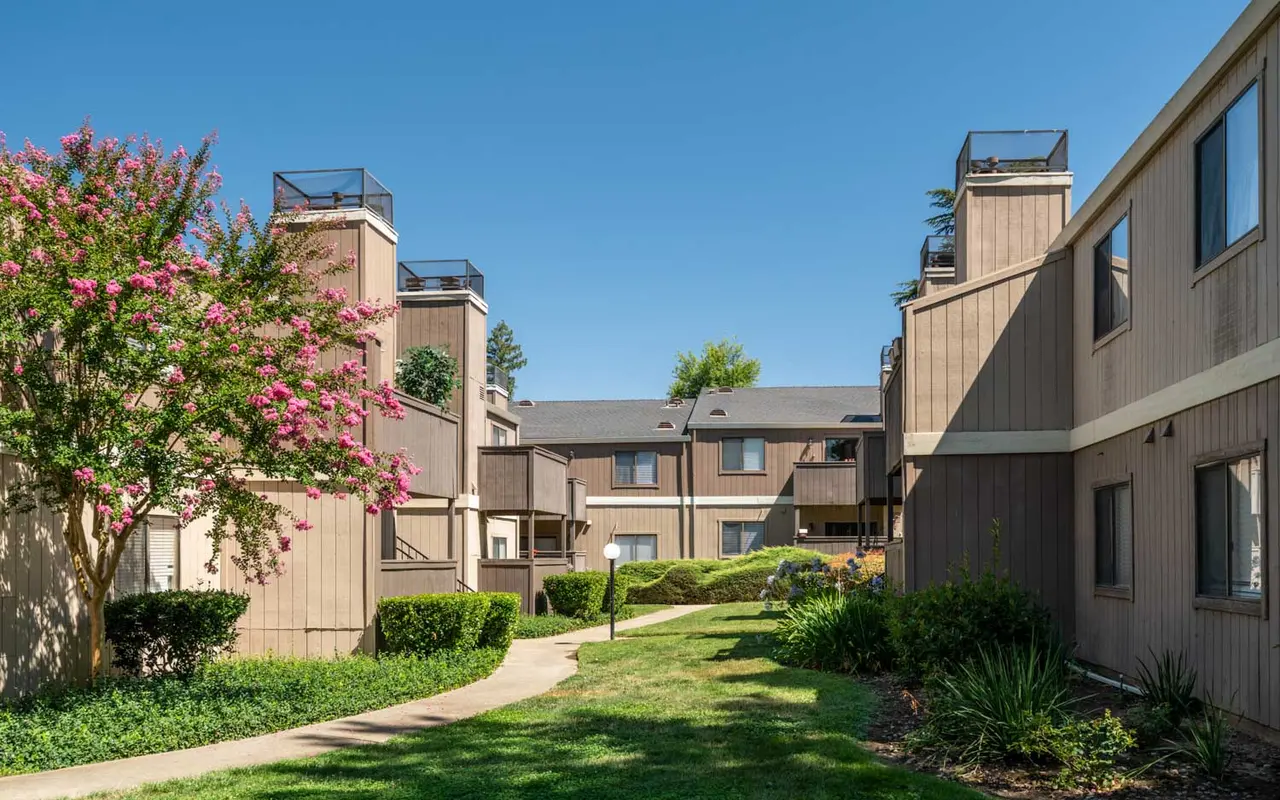 A courtyard view of an apartment complex featuring tan buildings, walkways, and green landscaping with flowering plants.