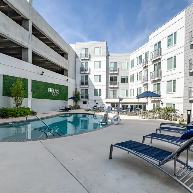 Apartment Complex Pool Area A modern apartment complex featuring a pool area surrounded by lounge chairs and greenery. The building has multiple levels and balconies, with a parking structure nearby.