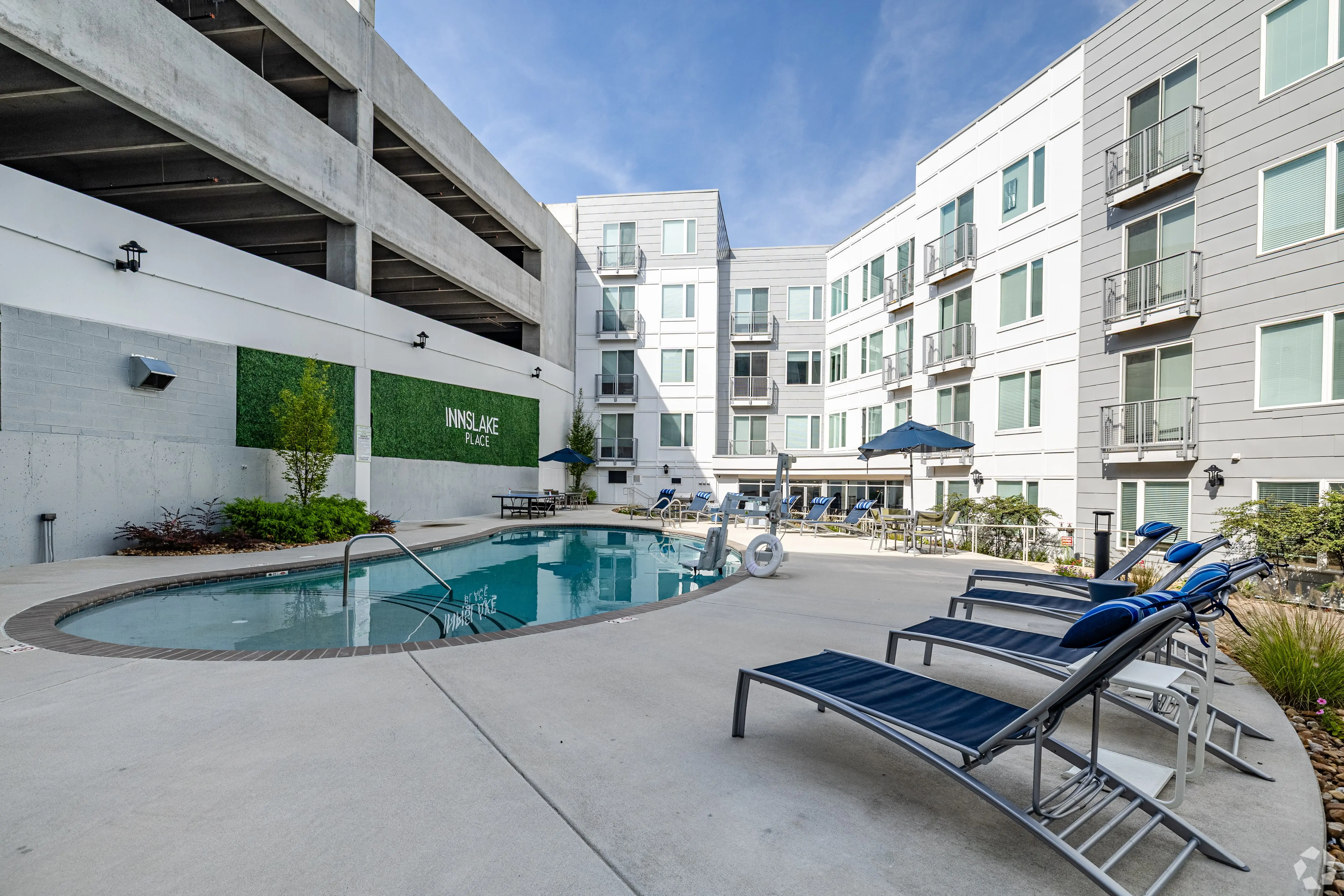A modern apartment complex featuring a pool area surrounded by lounge chairs and greenery. The building has multiple levels and balconies, with a parking structure nearby.