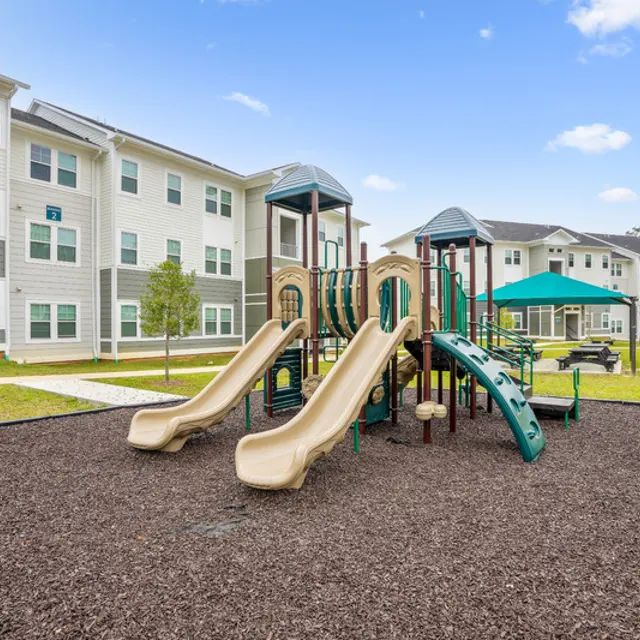 A children's playground with slides and climbing equipment, surrounded by grass and residential buildings under a blue sky.