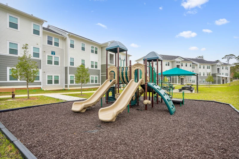 Playground with slides and climbing structure on a soft surface, surrounded by grass and residential buildings in the background.