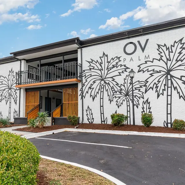 Exterior view of a two-story building with a mural of palm trees on its white walls. The building features an orange accent at the entrance and landscaping with bushes.