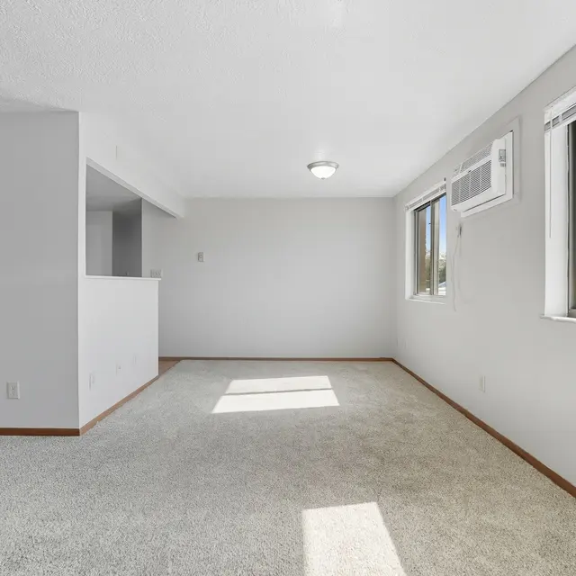 A light and airy living room with beige carpet, large windows allowing natural light, and a view into a kitchen area with a black refrigerator.