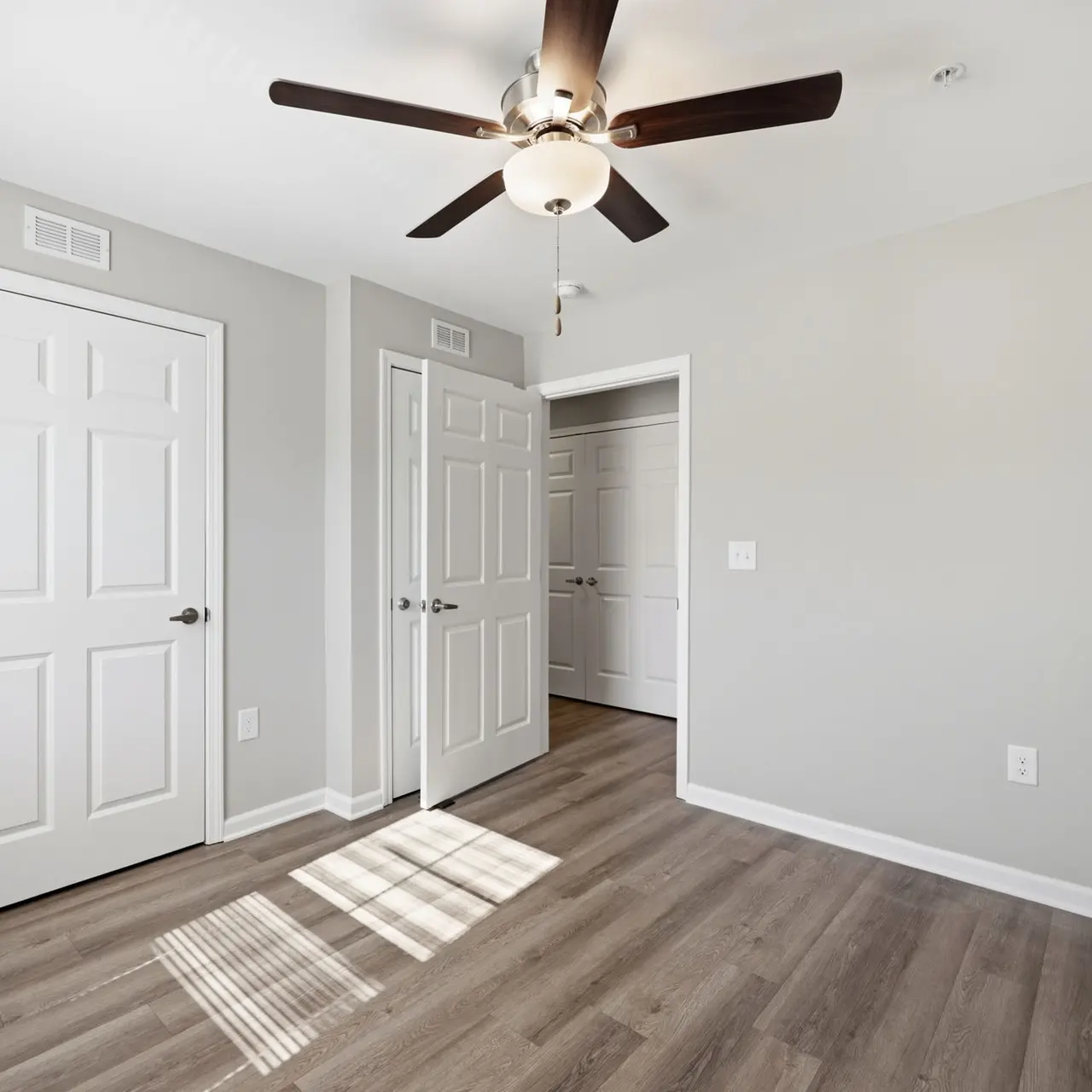 A modern bedroom featuring light grey walls, a ceiling fan, and two closed doors. The room has wooden flooring and shows sunlight casting shadows on the floor.