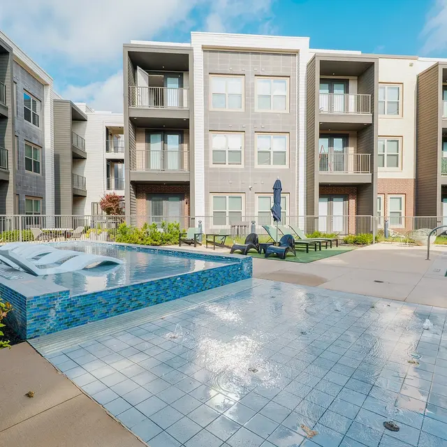 A modern apartment complex featuring a pool area with a spa and lounge chairs, surrounded by greenery.