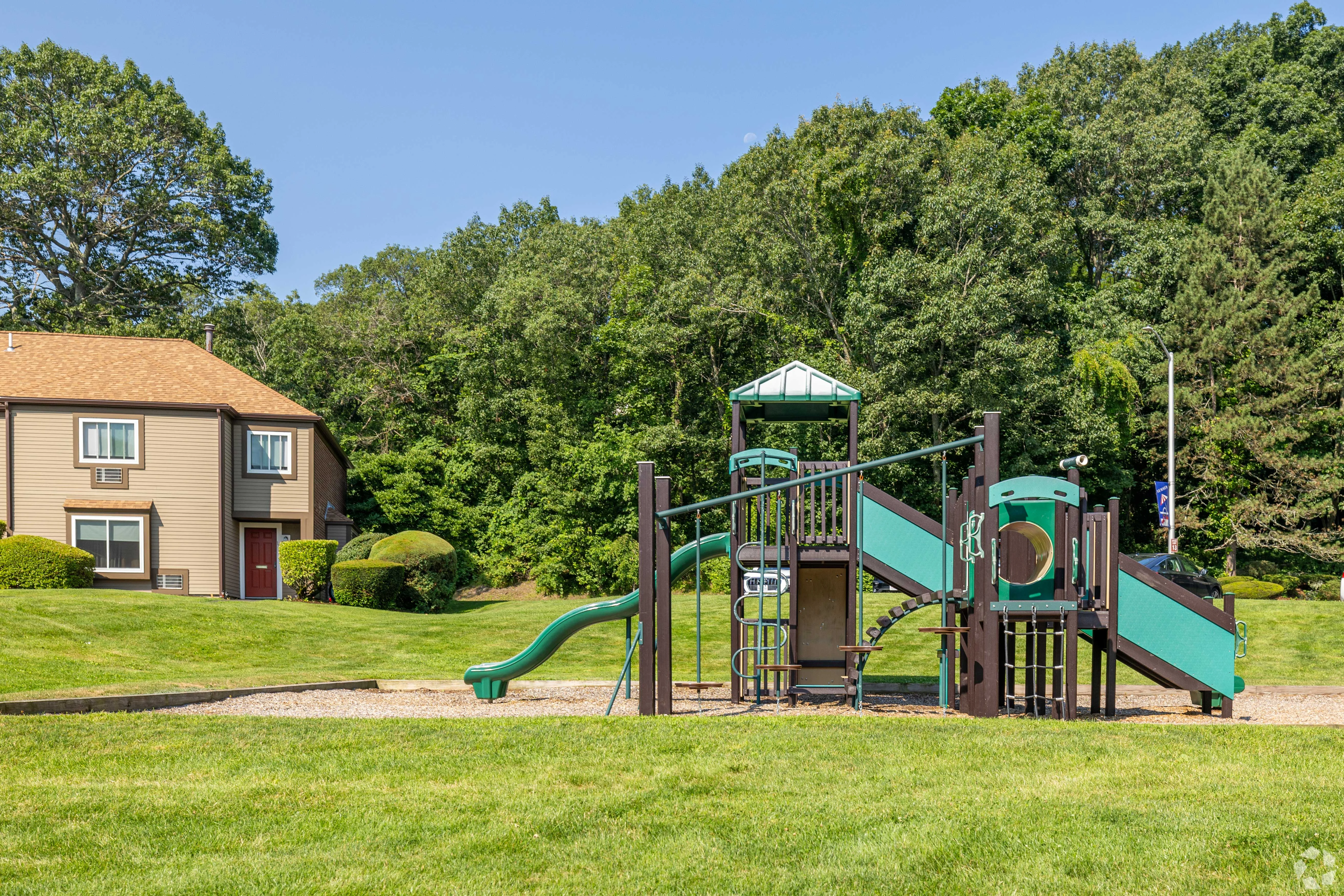 Playground in Residential Area A playground featuring slides and climbing structures, situated on a grassy area beside a residential building and surrounded by trees.