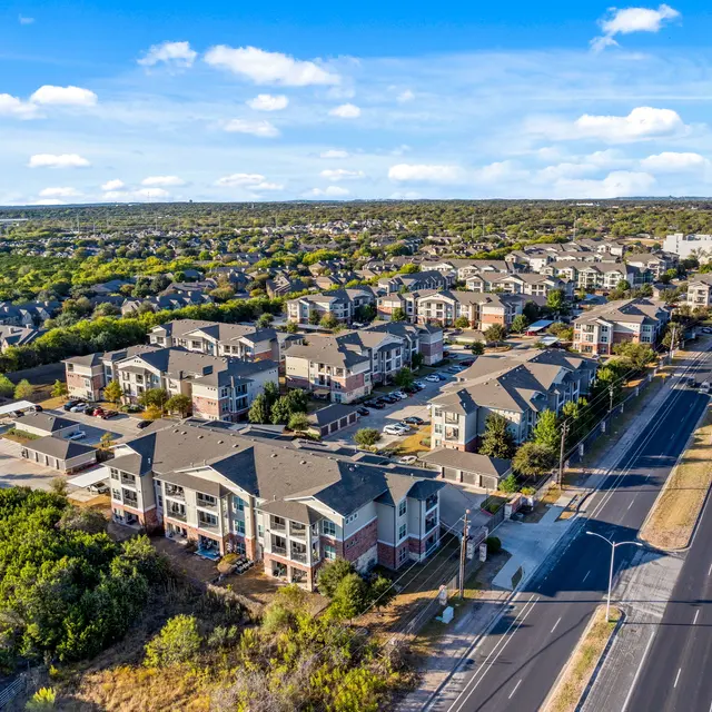Aerial view of a suburban apartment complex surrounded by greenery and a road running alongside.