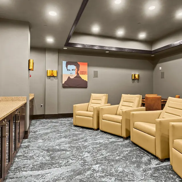 A cozy lounge area featuring comfortable beige chairs, a large flat-screen TV on the left, and a decorative wall art piece. The flooring is carpeted with a textured design, and subtle wall lighting enhances the ambiance.