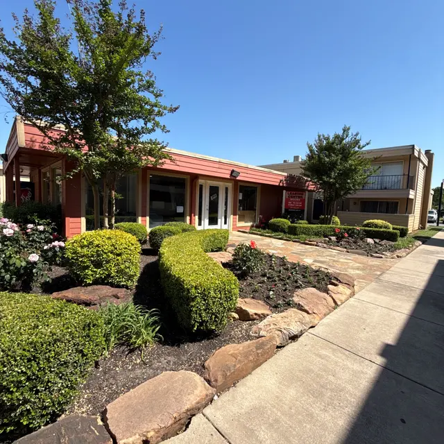 Exterior view of a building with a red roof and landscaped garden featuring shrubs and flowers, clear blue sky in the background.