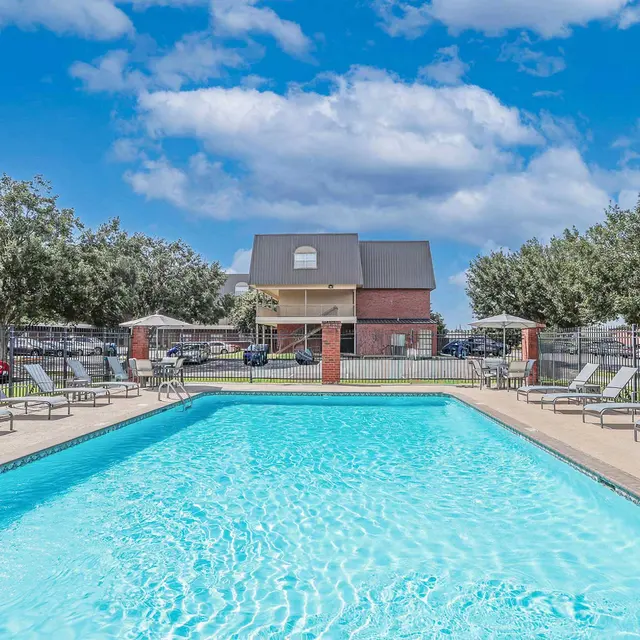 A swimming pool surrounded by lounge chairs and greenery, with a building in the background and a bright blue sky overhead.