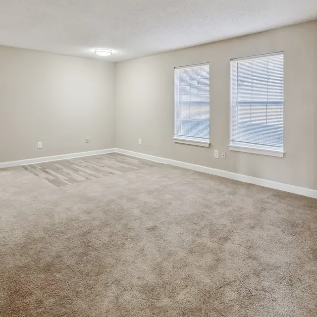 A spacious, empty living room with light beige walls and plush carpet. Two rectangular windows allow natural light, and a door is visible on the left leading to another area. The room is well-lit with a ceiling light fixture.