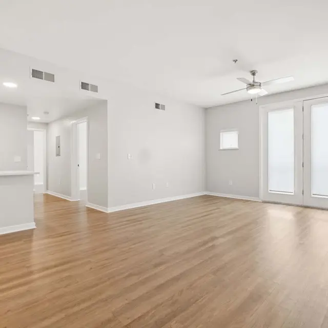 Modern Apartment Living Room A modern apartment living room featuring light hardwood floors, light grey walls, a ceiling fan, and large windows with doors that lead outside.