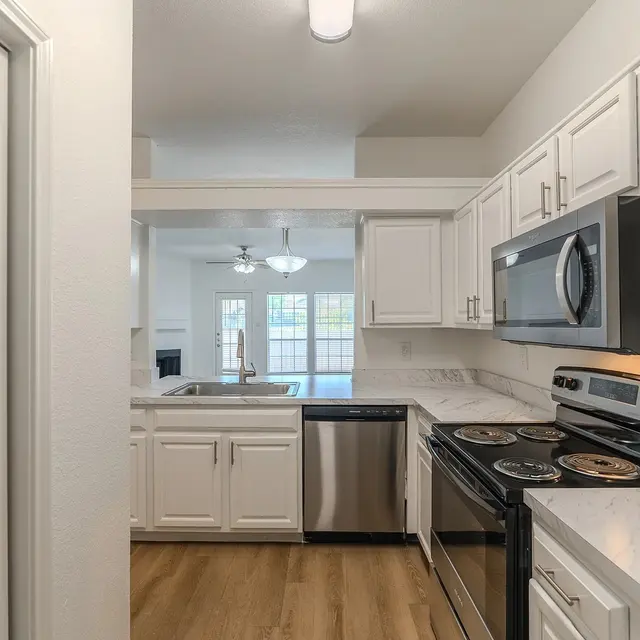 A modern kitchen with white cabinets, stainless steel appliances, and a marble countertop. The kitchen opens up to a living area visible in the background.