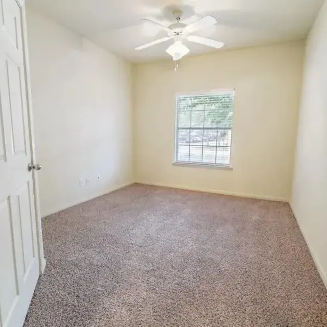 An empty room with beige walls and light carpet, featuring a ceiling fan and a window with a view outside.