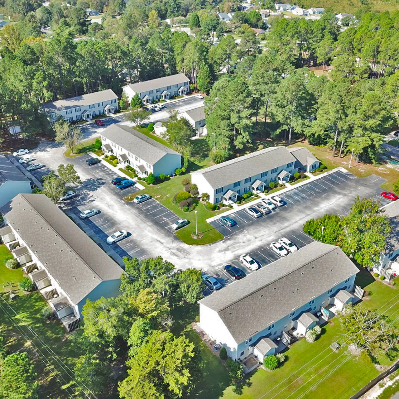 Aerial view of a multi-building apartment complex surrounded by trees, with parking lots and green areas visible.