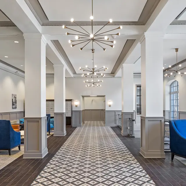 A modern hotel lobby featuring a long corridor with patterned flooring, tall white pillars, and contemporary light fixtures. Blue armchairs are arranged around the space, with framed black-and-white photographs on the walls.