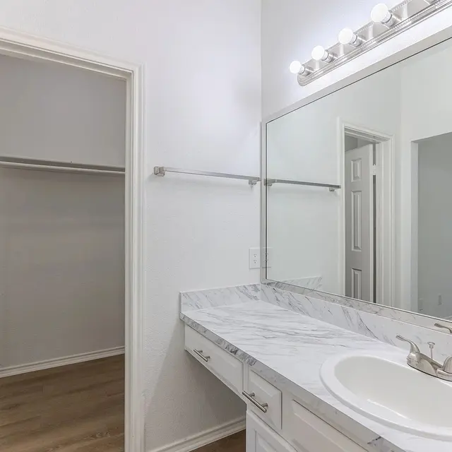 A modern bathroom featuring a marble countertop with a sink, large mirror, and light fixtures above. To the side, there is a walk-in closet, and the walls are painted white, complemented by wooden flooring.