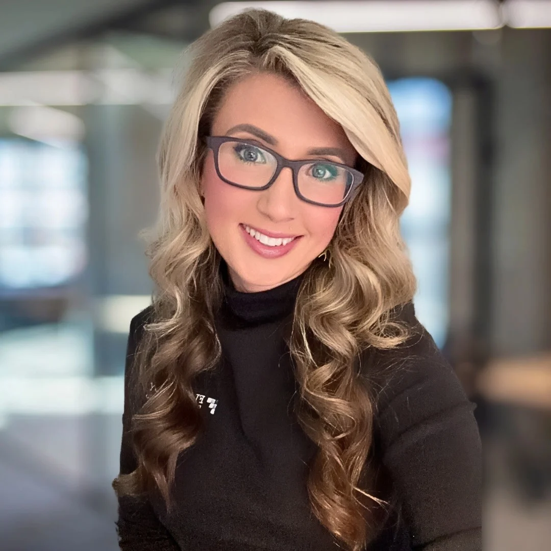 A woman with long, wavy blonde hair wearing glasses and a black top, smiling at the camera.