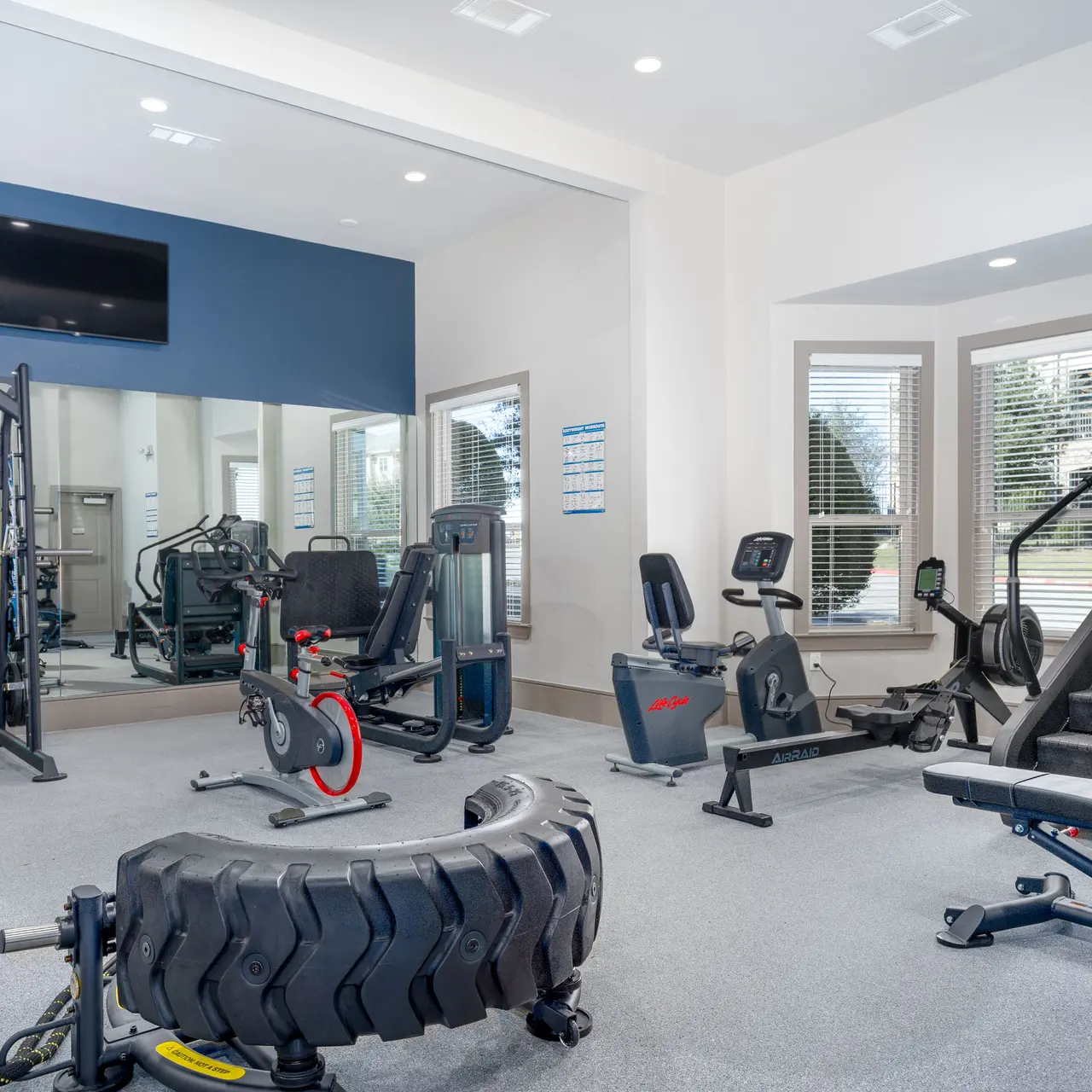 24/7 Fitness Center A modern gym interior featuring various exercise equipment including weight machines, a treadmill, and free weights, with large windows and mirrors providing a bright atmosphere.