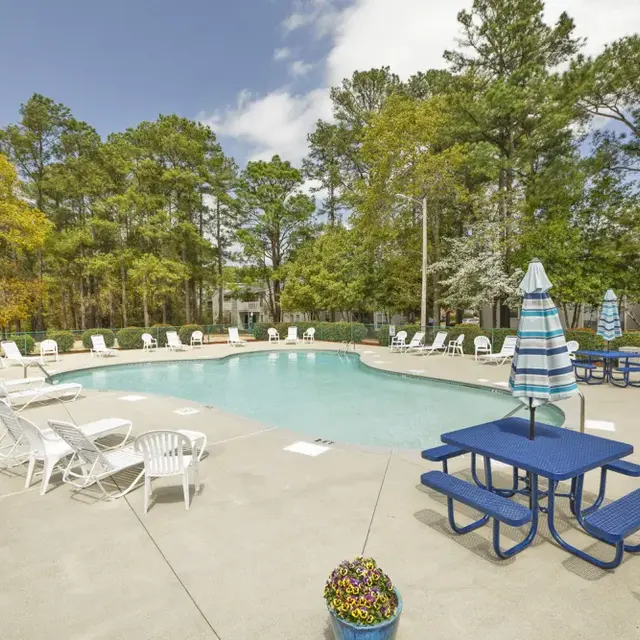A swimming pool surrounded by white lounge chairs and blue picnic tables, with trees and a clear sky in the background.