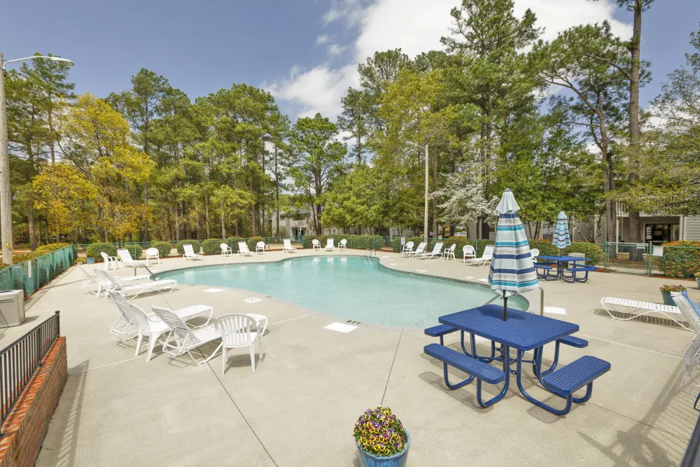 A swimming pool surrounded by white lounge chairs and blue picnic tables, with trees and a clear sky in the background.