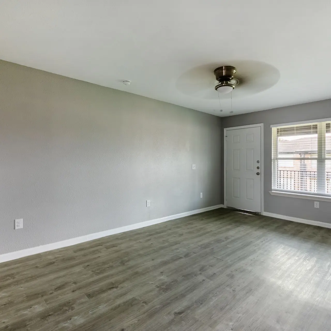 An empty living room featuring a gray wall, light wood flooring, and a ceiling fan. There is a door on the right side leading to the outside, and a window with blinds allowing natural light in.