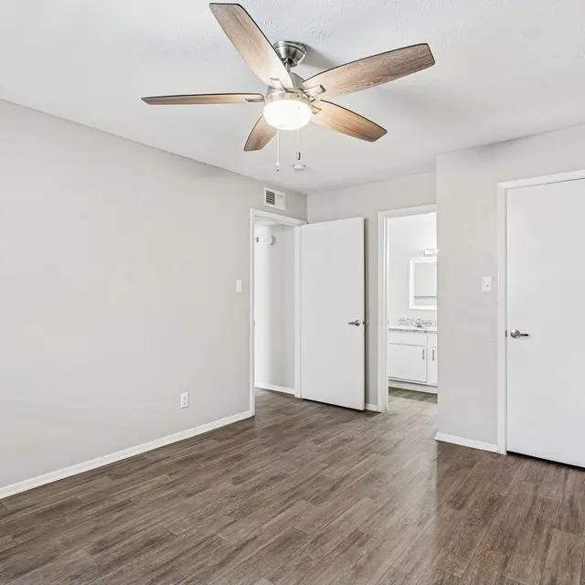 A bright and airy empty room with a ceiling fan, featuring beige walls and wood-style flooring. A doorway leads to a bathroom area, enhancing the spaciousness of the room.