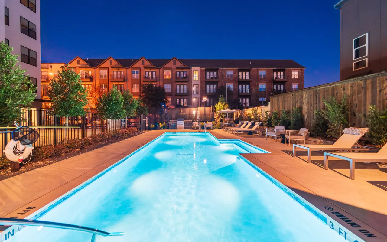 A well-lit swimming pool area at night surrounded by lounge chairs and trees, with an apartment building in the background.
