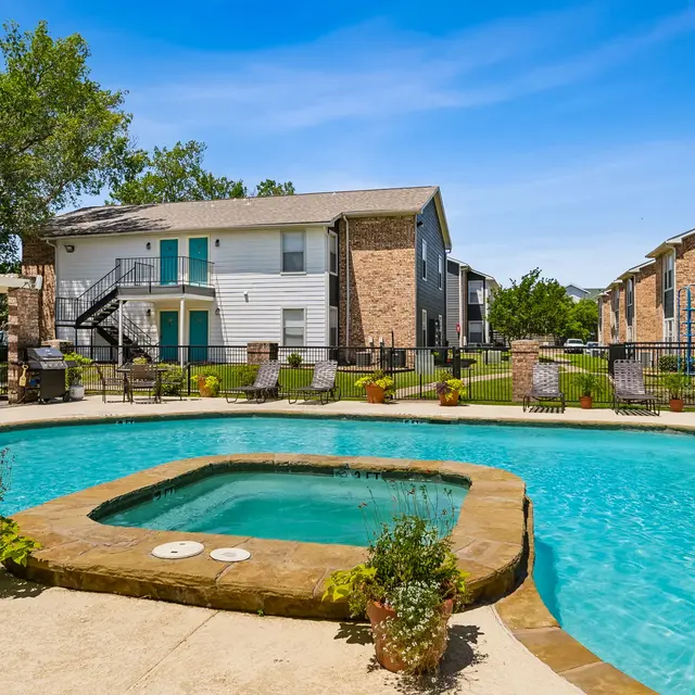 A serene pool area featuring a sparkling turquoise pool with a jacuzzi, surrounded by landscaped greenery and apartment buildings in the background under a clear blue sky.
