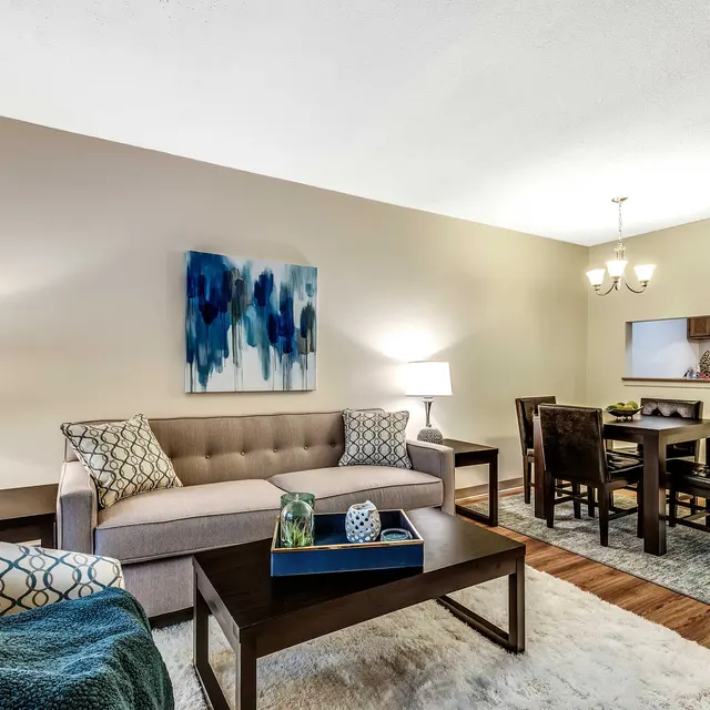 A stylish modern living room featuring a beige couch with decorative pillows, a blue patterned armchair, a wooden coffee table, and a dining area with a wooden table and chairs in the background. There is a large abstract painting on the wall and a soft rug on the floor.