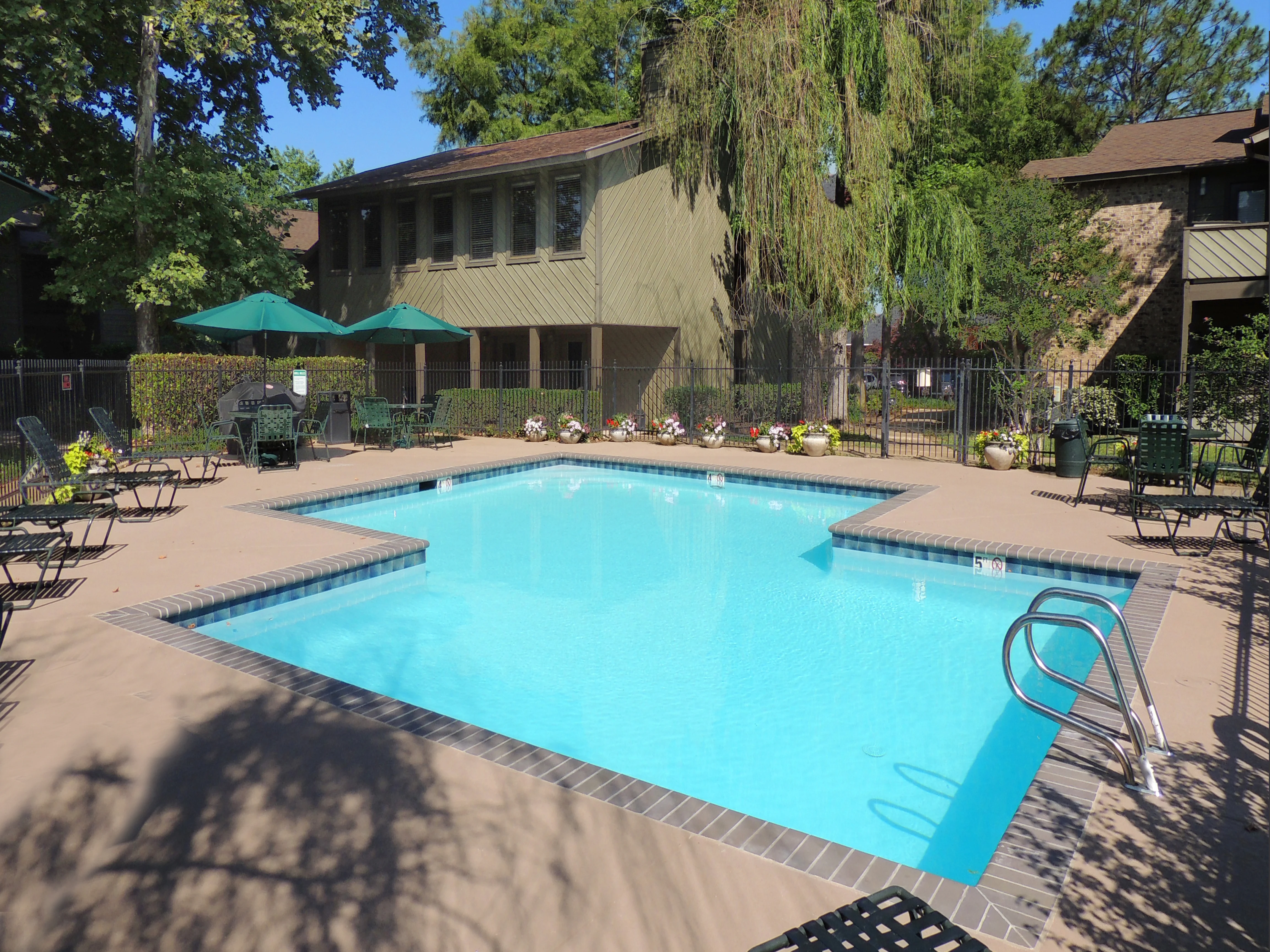 A clear swimming pool surrounded by lounge chairs, a fence, and lush greenery in an apartment complex.