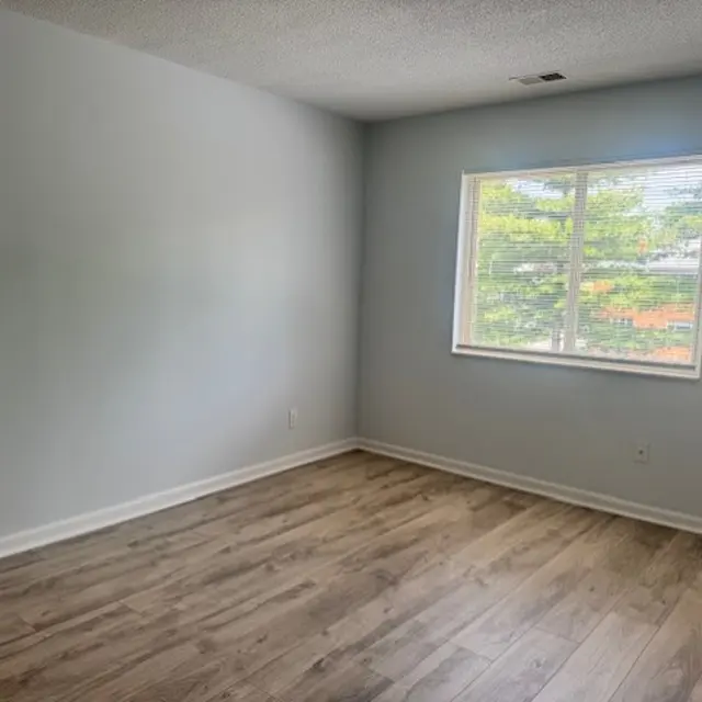 A vacant room with light gray walls and wooden flooring. A window on one wall allows natural light to enter, showcasing trees outside.