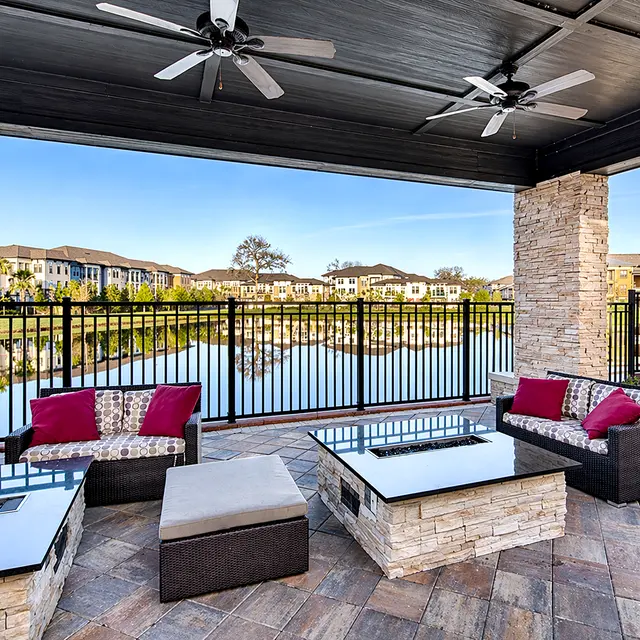 A stylish outdoor lounge area featuring dark wicker furniture with colorful cushions, set under a shaded patio with ceiling fans. The backdrop includes a serene view of a lake and residential buildings.